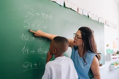 Teacher pointing at the chalkboard while talking to a student. Featured Image